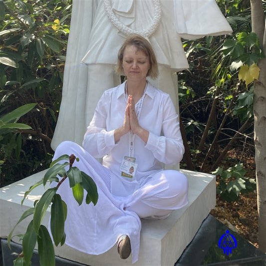 Eva Marquez in India, during a spiritual resort/training, sitting at the base of a white spiritual statue with her back to the statue. She sits with her eyes closed and her legs crossed in a meditating position. Her hands are held in a praying position in front of her chest, and she is wearing sandals. The area is a very peaceful and tranquil wooded area with many plants and trees surrounding her and the statue. Eva's Logo is imprinted on the lower right side of photo.