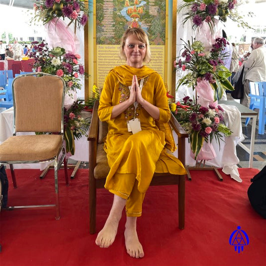 Eva Marquez in India, during a spiritual resort/training, sitting in a chair, left leg over right leg, in front of table full of flowers with an Indian spiritual picture in the center. She is bare feet and has her hands in a praying position in front of her chest and is wearing gold colored Traditional Sacred Indian Mediation Garb. Eva's Logo is imprinted on the lower right side of photo.